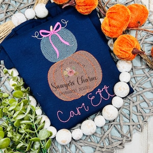 May include: Navy blue long-sleeve shirt with embroidered pumpkins and the text "Sawyer's Charm" and "Carrett". The top pumpkin has a pink bow. The shirt is surrounded by white beads and orange pumpkins.