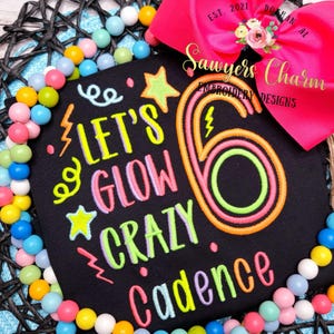 May include: A black embroidered patch with the text "LET'S GLOW CRAZY Cadence" and a large number "6". The design includes stars, lightning bolts, and swirls in neon colors. A bright pink bow and colorful beads surround the patch.
