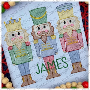 May include: Three embroidered nutcracker figures, one in green, one in blue, and one in red, with the name "JAMES" embroidered below them.