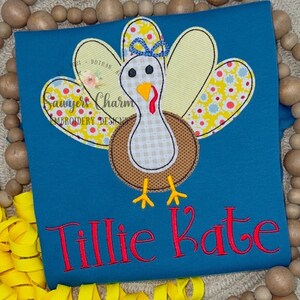 May include: A teal blue t-shirt with an embroidered appliqué of a Thanksgiving turkey. The turkey is brown, white and yellow with a blue bow. The t-shirt also has the name "Tillie Kate" embroidered in red.