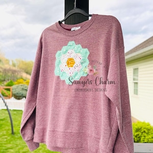 May include: A maroon sweatshirt with a floral design. The design is a light blue and white flower with a yellow center. The text "Sawyers Charm Embroidery Designs" is below the flower.