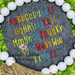 May include: A gray felt square embroidered with the alphabet in rainbow colors. The letters are in both uppercase and lowercase. The text "Sawyers Charm Embroidery Designs" is embroidered at the bottom.