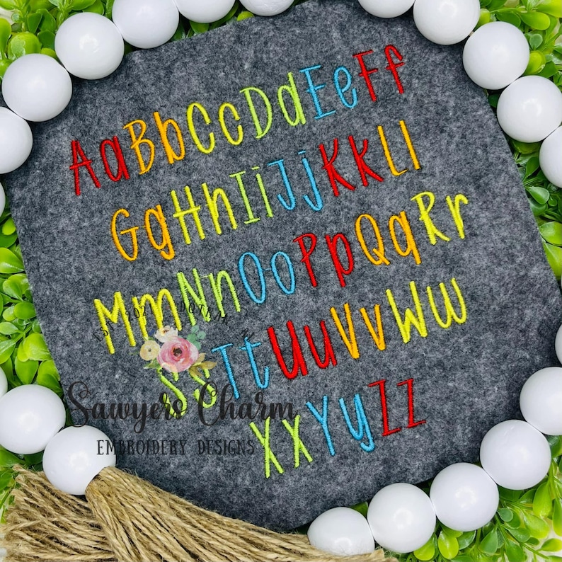 May include: A gray felt square embroidered with the alphabet in rainbow colors. The letters are in both uppercase and lowercase. The text "Sawyers Charm Embroidery Designs" is embroidered at the bottom.