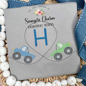Puede incluir: Una camiseta gris con camiones monstruo bordados, la letra "H" en un corazón y el texto "Sawyers Charm Embroidery Designs" y "Est. 2021 Dothan, AL".