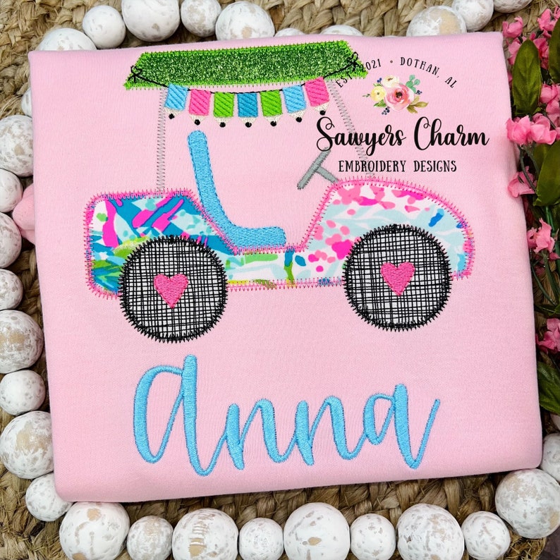 May include: Pink embroidered t-shirt with a colorful jeep design and the name "Anna" embroidered in blue. The jeep has a green canopy and pink and blue floral accents. The text "Sawyers Charm Embroidery Designs" is also embroidered on the shirt.