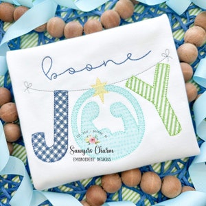 May include: White onesie with a blue, green, and white embroidered "JOY" banner with a star and a nativity scene in the center. The onesie also has the name "Boone" embroidered above the banner. The text "Est. 2021 - Dotted Air - Sawyers Charm Embroidery Designs" is embroidered below the banner.