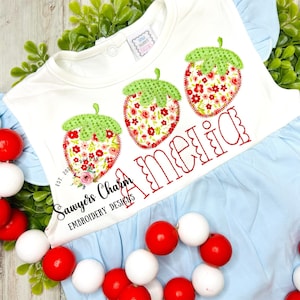 May include: A white baby onesie with a blue ruffle trim. The onesie has three embroidered strawberries with green leaves and red and pink floral patterns. The name "Amelia" is embroidered in a cursive font below the strawberries. The text "Sawyers Charm Embroidery Designs" is embroidered on the bottom left corner of the onesie.