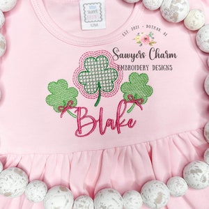 May include: A light pink baby dress with a ruffled hem, featuring an embroidered shamrock design in green and pink. The name "Blake" is embroidered in pink script. The dress has a tag that says "12M". The text "Sawyers Charm Embroidery Designs" is also visible.