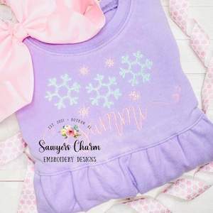 May include: Purple baby onesie with a pink and white bow on top. The onesie has a design of snowflakes and the name "Sammy" embroidered in pink. The text "Sawyers Charm Embroidery Designs" is also embroidered on the onesie.