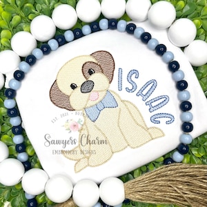 Puede incluir: Un babero blanco bordado con un cachorro de dibujos animados con una pajarita azul y el nombre "Isaac" bordado en azul. El babero está rodeado por una guirnalda decorativa de cuentas blancas y azules y vegetación.