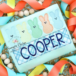 May include: A light blue t-shirt with a blue and white plaid banner that says "COOPER" in blue embroidery. Above the banner are four embroidered pastel-colored Peeps, a yellow Peep, a green Peep, an orange Peep, and a blue Peep.