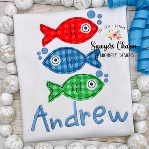 May include: A white t-shirt with three embroidered fish in red, blue, and green. The fish have a checkered pattern and black outlines. The name "Andrew" is embroidered in blue below the fish. The text "Sawyers Charm Embroidery Designs" is embroidered in black on the top right corner of the shirt.