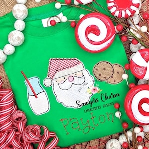 May include: Green t-shirt with embroidered images of Santa Claus, a glass of milk with a straw, and two cookies. The text "Sawyers Charm Embroidery Designs" and "Payton" are also embroidered on the shirt.