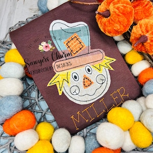 May include: Brown t-shirt with an embroidered scarecrow face and the text "Sawyers Charm Embroidery Designs" and "Miller".