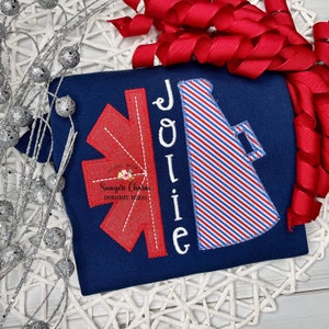 May include: Navy blue t-shirt with a red and white star of life design on the left and a red, white, and blue striped megaphone on the right. The name "Jolie" is embroidered in white. The shirt is decorated with a red ribbon.
