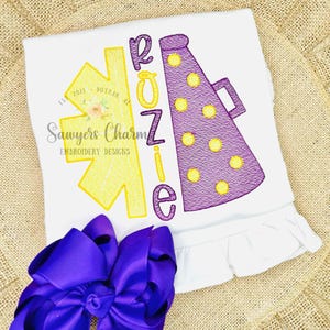 May include: White t-shirt with embroidered yellow and purple cheerleading design. The design includes a megaphone, a starburst, and the name "ROZIE". A large purple bow is at the bottom of the shirt.