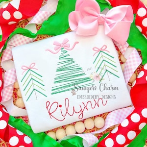 May include: White embroidered shirt featuring three green Christmas trees, each adorned with a pink bow. The shirt also includes the name "RiLynn" embroidered in red cursive lettering. The text "Sawyers Charm Embroidery Designs" is also embroidered on the shirt.