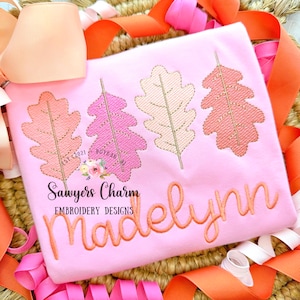 May include: Pink children's shirt with a design of three embroidered autumn leaves in shades of pink, peach, and cream. The text "Sawyers Charm Embroidery Designs" and the name "Madelynn" are also embroidered on the shirt.
