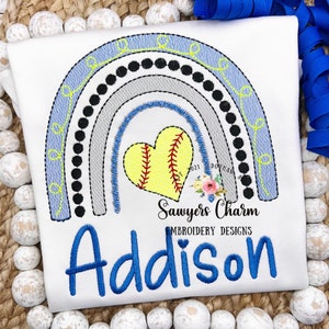 May include: A white t-shirt with a rainbow design featuring a softball in a heart shape. The text "Sawyers Charm Embroidery Designs" and the name "Addison" are embroidered on the shirt.