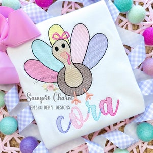 May include: A white babygrow with a colourful embroidered turkey and the name "Cora" in rainbow lettering. The text "Sawyers Charm Embroidery Designs" is also embroidered on the babygrow.