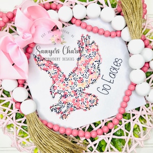 Peut inclure: Une couronne rose et blanche avec un motif d'aigle floral. La couronne comporte des perles roses, un nœud rose et une gland en jute. L'aigle est rempli d'un motif floral et des mots "Go Eagles".