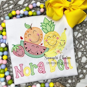 May include: A white t-shirt with embroidered fruit designs, including a watermelon, pineapple, banana, orange, and strawberry. The text "Nora Bell" is embroidered in pink, yellow, and green letters below the fruit. The text "Sawyers Charm Embroidery Designs" is embroidered in black below the name.