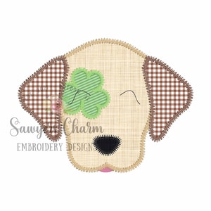 May include: A stitched dog face with a shamrock over one eye. The dog's ears are brown and white checkered, and the shamrock is green and white striped. The dog has a black nose and a pink tongue. The text "Sawyer's Charm Embroidery Design" is in the lower left.