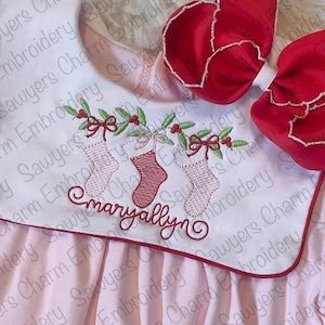 May include: A pink baby dress with a red trim and embroidered Christmas stockings with holly and berries. The name "Maryallyn" is embroidered on the dress.