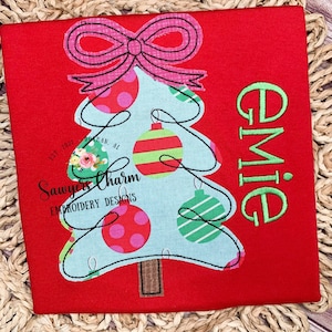 May include: An embroidered Christmas tree in red with a pink bow, green and red baubles, and the name "Emie" embroidered in green on a red background.