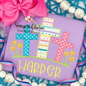 May include: A lavender long-sleeve shirt with three embroidered crosses in blue, yellow, and pink, each with a unique pattern. The name "Harper" is embroidered in yellow script. The shirt is decorated with yellow flowers and green grass.