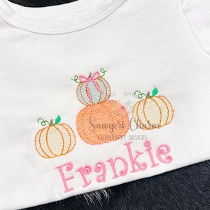 May include: White t-shirt featuring embroidered pumpkins in orange, light orange, and light blue, with green vines and a pink bow. The name "Frankie" is embroidered in pink script below the pumpkins. "Sawyer Charm Embroidery Designs" is also embroidered.