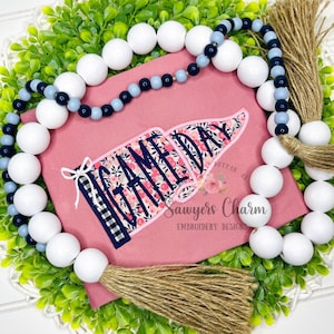 May include: A pink rectangular sign with the text "GAMEDAY" in a floral design, accented with a navy blue and white plaid pattern. The sign is surrounded by a white beaded garland with navy and light blue beads, and a green leafy base.