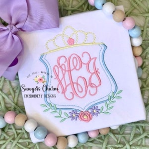 May include: A white embroidered shirt with a pink monogram in a blue and white shield with a crown above it. The monogram is "HCG". The shirt has a floral design below the shield. The text "Sawyers Charm Embroidery Designs" is below the shield.