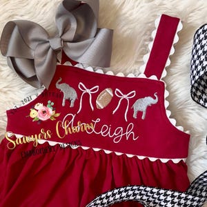 May include: A red romper with white scalloped trim, featuring embroidered elephants, bows, and a football. The romper has the name "Charms Leigh" embroidered in gold script, with a large gray bow and a black and white houndstooth ribbon.