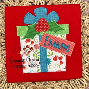 May include: A red, green, and blue embroidered applique of a Christmas present with a white tag that says "EMMIE". The present is decorated with a green bow and a red flower. The applique is on a red fabric background.