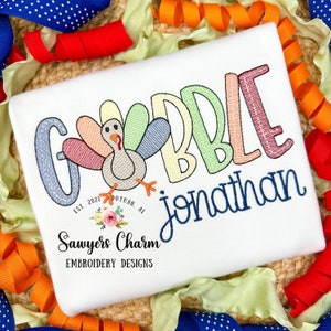 May include: A white t-shirt with a colorful embroidered design that says "Gobble" with a turkey in the middle. The name "Jonathan" is embroidered below the design. The text "Sawyers Charm Embroidery Designs" is embroidered below the name.