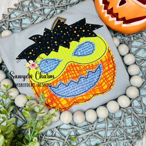 Pode incluir: Uma camiseta cinza com um design de abóbora bordado. A abóbora é amarela, laranja e azul com olhos pretos e um sorriso largo. O texto "Sawyers Charm Embroidery Designs" é bordado na camiseta.