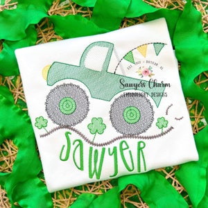 May include: Green monster truck with a yellow light and a green, yellow, and white flag in the back. The truck is driving on a brown path with green shamrocks. The name "Sawyer" is written in green below the truck. The text "Sawyers Charm Embroidery Designs" is written in black below the truck.