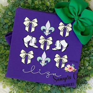 May include: Purple t-shirt with embroidered white bows, fleur-de-lis, and boots. The shirt has the text "Sawyers Charm Embroidery Designs" and "Dothan, AL" on it.