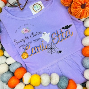 May include: Purple  long-sleeved shirt with a  Halloween-themed embroidery design. The design includes a ghost, spider web, spider, pumpkin, and the text "Sawyers Charm Embroidery Designs" and "Carli Etta".
