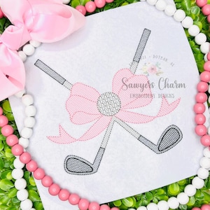May include: Embroidered design of two crossed golf clubs with grey club heads and handles, and a pink bow with a golf ball centre. The design is on a white background, surrounded by pink and white beads and greenery. Text reads "Sawyers Charm Embroidery Designs".