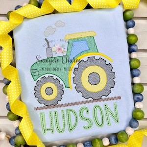 May include: A light blue t-shirt with an embroidered green tractor with a yellow wheel and the name "HUDSON" in green letters. The text "EST 2021 - DOTHAN" is embroidered above the tractor.