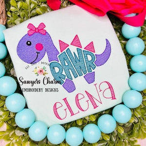 May include: A light blue t-shirt with a purple dinosaur with a pink bow and the word "RAWR" in teal. The dinosaur is holding a pink flower. The text "Sawyers Charm Embroidery Designs" and "Elena" are also embroidered on the shirt.