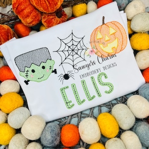 May include: White embroidered t-shirt with a green Frankenstein, a spider web with a spider, and a jack-o-lantern. The name "ELLIS" is embroidered in green below the design. "Sawyers Charm Embroidery Designs" is embroidered in black below the jack-o-lantern.