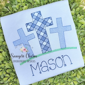 May include: A light blue baby onesie with a blue embroidered cross design and the name "Mason" embroidered below. The cross is a plaid pattern and there is a green embroidered line below the cross.