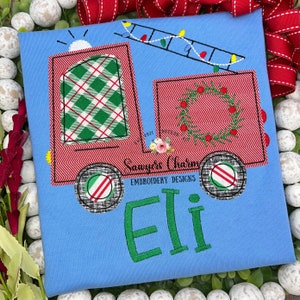 May include: A blue t-shirt with an embroidered design of a red fire truck with a green and white checkered window, a wreath on the back, and a ladder with lights. The truck has black wheels with red and white stripes. The name "Eli" is embroidered in green below the truck. The text "Sawyer's Charm Embroidery Designs" is also embroidered on the shirt.