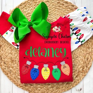 May include: Red shirt with the name "Delaney" embroidered in green cursive lettering.  Five colorful light bulbs are embroidered below the name, with a string of lights connecting them.  The shirt has the text "Sawyers Charm Embroidery Designs" on the front.