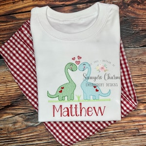 May include: White t-shirt with red and green embroidered dinosaurs holding hearts. The shirt also has the text "Sawyers Charm Embroidery Designs" and "Matthew" embroidered on it.