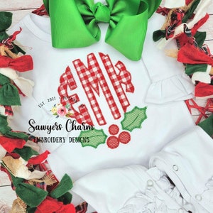 May include: White baby onesie with a red and white buffalo plaid monogram "GMR" with green holly leaves and red berries. The onesie has a ruffled sleeve and the text "Sawyers Charm Embroidery Designs" is printed below the monogram.