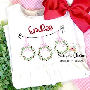 以下が含まれることがあります： 赤い刺繍で「Emree」と刺繍された白いベビーロンパース。ピンクのリースが3つ、緑の葉と赤いベリーが飾られ、紐で吊り下げられています。ロンパースには「Sawyers Charm Embroidery Designs」と「Est. 2021 - Dothan, AL」の文字も刺繍されています。
